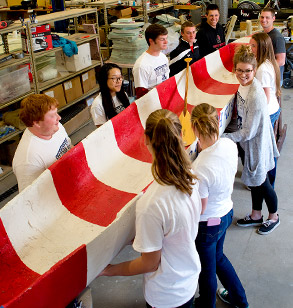 Several students lift a red and white striped concrete canoe.