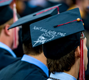 Image shows the back of the heads of three male graduating students during Commencement. They are wearing black graduation gowns and caps.