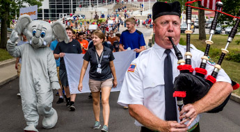 image shows a man playing bagpipes at the front of a long march of freshmen as they are led by a resident assistant and the mascot, Rosie, up the road from the Sports and Recreation Center to the main part of campus during orientation.
