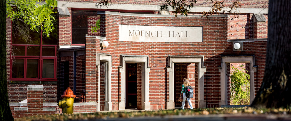 Image shows view looking into the courtyard outside Hadley, Moench, and Olin halls. Image shows a female student walking through a majestic brick entrance into Moench Hall. 
